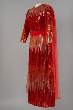 A long-sleeved, floor-length red gown with gold sequins in vertical patterns and a matching red sash at the waist. The dress features a sheer red cape attached at the shoulders, draping down the back. The gown is displayed on a mannequin.