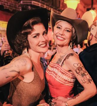 Two smiling women wearing cowboy hats and sleeveless outfits pose together at a lively indoor event. Both have tattoos on their arms and appear happy, with warm lighting and people visible in the background.