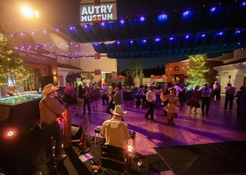 A live band performs on stage while people in cowboy hats dance under string lights at an outdoor evening event at the Autry Museum. The venue is lively with festive lighting and a relaxed atmosphere.