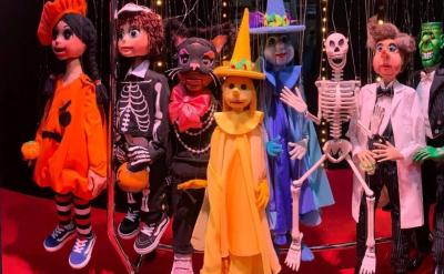 Seven marionette puppets dressed in colorful Halloween costumes, including a pumpkin, skeleton, cat, witch, and Frankenstein, stand together on a red floor with string lights in the background.