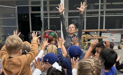A group of young children sit outdoors with their hands raised, mirroring an adult who is leading them in an activity. They are gathered near benches and a fence, possibly at a school playground.