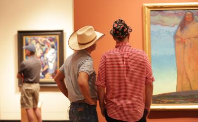 Two men, one in a cowboy hat and another in a floral-patterned cap, stand side by side viewing a framed painting in an art gallery. Another person in casual clothes looks at a different painting in the background.