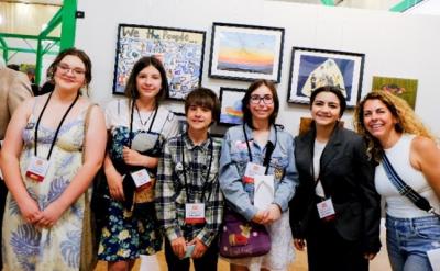 A group of six people, including five young students and one adult, stand smiling in front of an art display featuring colorful paintings and a collage reading "We the People." All wear nametags and casual to semi-formal attire.