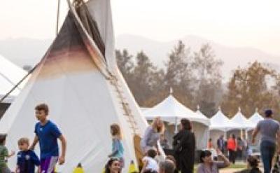 People, including families and children, gather around a large tipi and several white tents at an outdoor event on a sunny day, with trees and mountains visible in the background.