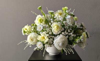 A white ceramic vase filled with white and pale yellow flowers, including roses and daisies, sits on a black table against a plain gray background.