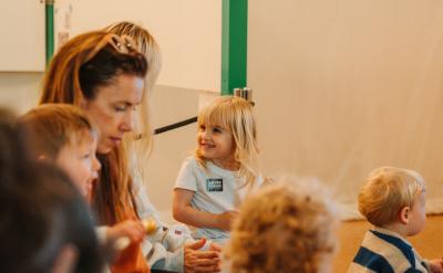 A group of young children sit together indoors, with one blonde girl smiling and looking at an adult nearby. The room has soft lighting and a relaxed atmosphere.