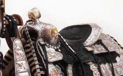 An ornate, Western-style saddle with detailed silver and gold embellishments, intricate leather tooling, and decorative straps, displayed against a plain white background.
