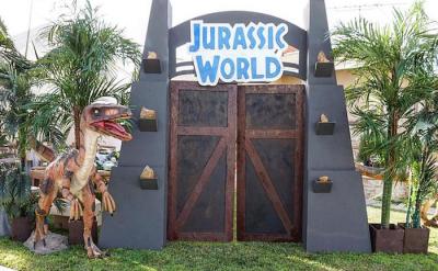 A Jurassic World-themed entrance with large double doors, palm trees, and a dinosaur statue beside the gate, set outdoors on a sidewalk.