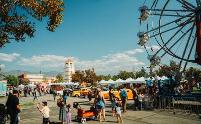 People enjoy a fair with a Ferris wheel, vintage cars, vendor tents, and a clock tower in the background under a sunny, blue sky. Families and children walk around the lively outdoor event.
