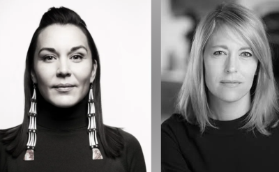 Black and white photo of two women in dark tops; the woman on the left wears long beaded earrings, while the woman on the right has shoulder-length hair. Both face the camera with neutral expressions.