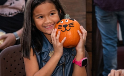 Smiling girl with long brown hair holds a small pumpkin decorated with a cat face and googly eyes. She wears a blue vest and a pink watch.
