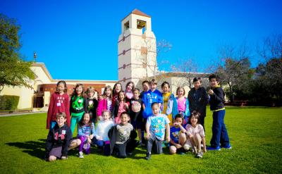 A group of elementary school-aged children posing for a photo on a grassy lawn in front of a mission-style building.