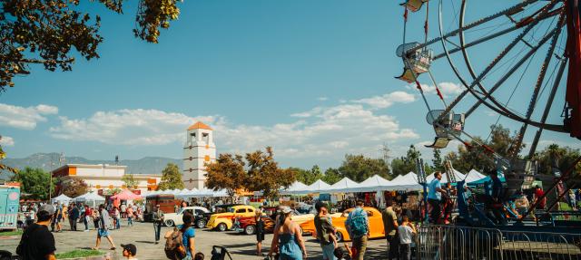 People enjoy a lively outdoor fair with a Ferris wheel, classic cars, vendor tents, and trees under a sunny blue sky. Families and children walk around, and a clock tower is visible in the background.