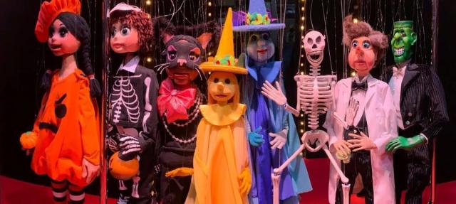 Seven marionette puppets dressed in colorful Halloween costumes, including a pumpkin, skeleton, cat, witch, and Frankenstein, stand together on a red floor with string lights in the background.