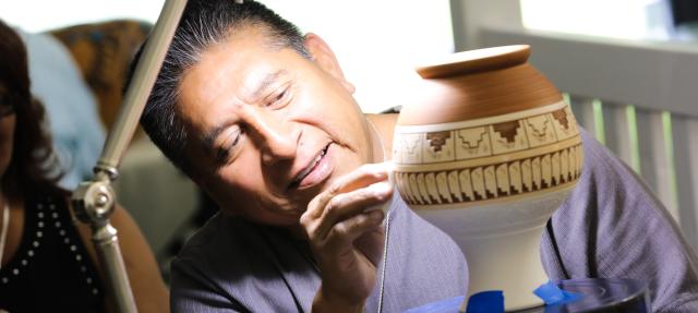 A man smiles while carefully painting intricate designs on a ceramic pot under a desk lamp, focusing on his detailed artwork.