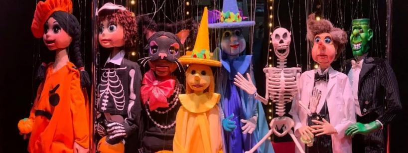 Seven marionette puppets dressed in colorful Halloween costumes, including a pumpkin, skeleton, cat, witch, and Frankenstein, stand together on a red floor with string lights in the background.