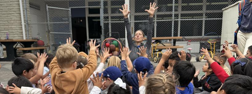A group of young children sit outdoors with their hands raised, mirroring an adult who is leading them in an activity. They are gathered near benches and a fence, possibly at a school playground.