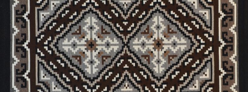 A rectangular rug with a geometric pattern in black, white, gray, and brown tones, featuring symmetrical diamond shapes and angular borders.