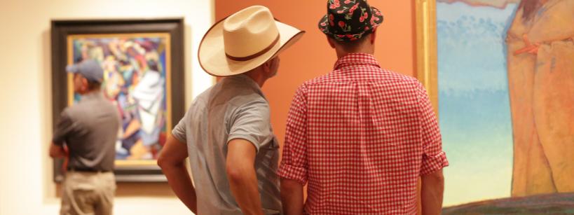 Two men, one in a cowboy hat and another in a floral-patterned cap, stand side by side viewing a framed painting in an art gallery. Another person in casual clothes looks at a different painting in the background.
