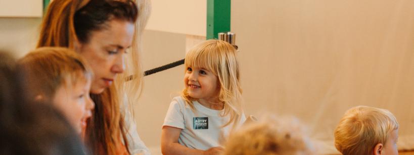 A group of young children sit together indoors, with one blonde girl smiling and looking at an adult nearby. The room has soft lighting and a relaxed atmosphere.