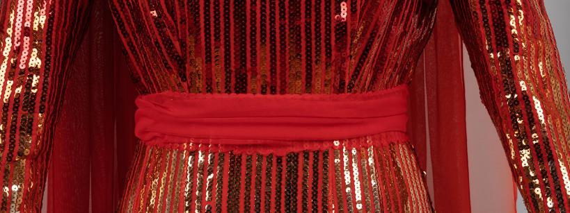 Close-up of a garment with vertical red and gold sequin stripes and sheer red fabric accents; features long sleeves and a solid red waistband or sash at the midsection.