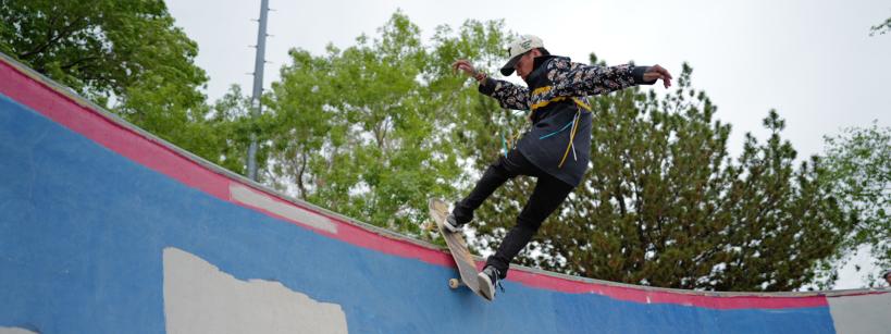 A skateboarder wearing a patterned shirt, black pants, and a white helmet performs a trick on the curved edge of a colorful skatepark ramp, with trees and a cloudy sky in the background.