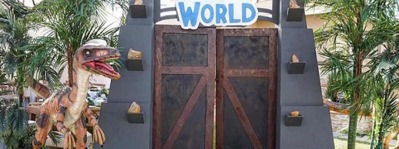 A Jurassic World-themed entrance with large double doors, palm trees, and a dinosaur statue beside the gate, set outdoors on a sidewalk.