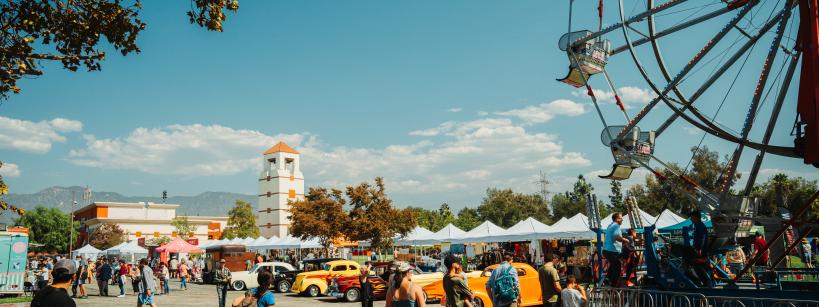 People enjoy a fair with a Ferris wheel, vintage cars, vendor tents, and a clock tower in the background under a sunny, blue sky. Families and children walk around the lively outdoor event.