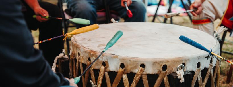 Four people sit around a large traditional drum, each holding a colored mallet and striking the drum together. The drum has a taut, laced surface and is played outdoors.
