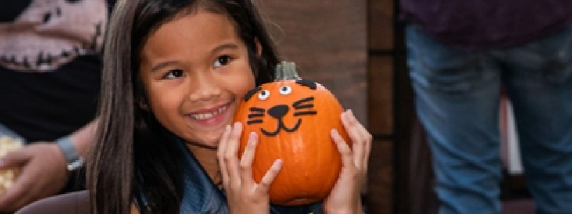 Smiling girl with long brown hair holds a small pumpkin decorated with a cat face and googly eyes. She wears a blue vest and a pink watch.