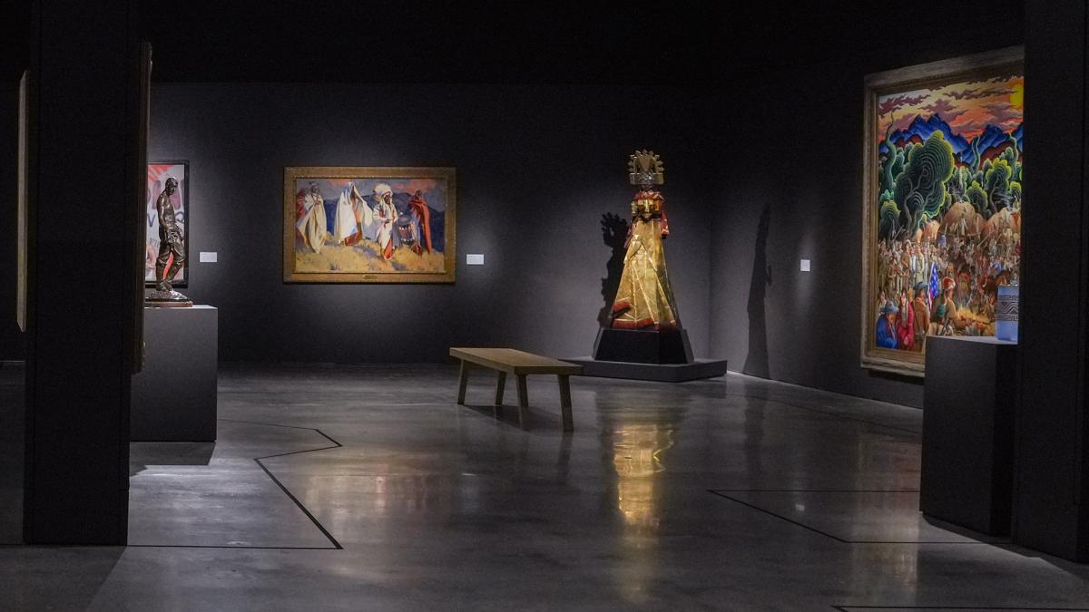 A dimly lit art gallery features colorful paintings on dark walls and a sculpture wearing a golden costume with a headdress. A wooden bench is placed on a polished concrete floor, providing a spot for visitors to sit and admire the artwork.