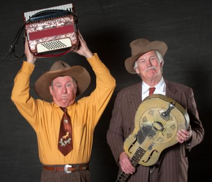 Two elderly men in cowboy hats pose for a photograph. The man on the left wears a yellow shirt and lifts an accordion over his head, making a surprised face. The man on the right wears a brown pinstripe suit and holds a guitar, smiling slightly.