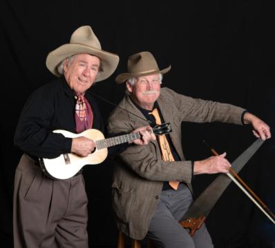 Two elderly men in cowboy hats smiling. One man is holding a small guitar, possibly a ukulele, and the other is playing a large hand saw with a bow. They are dressed in western-style clothing and are posing against a plain black background.