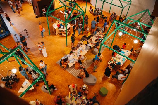 A busy indoor event with people gathered around tables covered in assorted materials. Green metal frames hang above, and various activities are happening throughout the space. Attendees include families and individuals in a creative workshop setting.