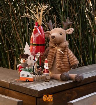 A display of festive holiday decorations including a plush reindeer, small snowman, Santa figurine, miniature camper, and trees. They sit on a wooden surface with tall grass in the background. The image is from the Autry Museum Store.