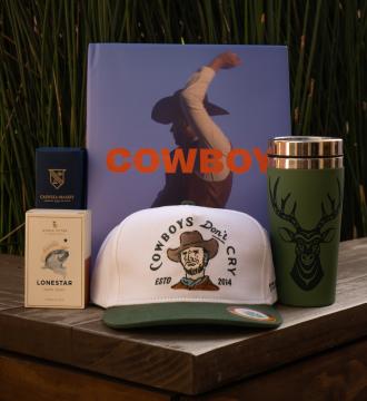 A collection of items on a wooden surface: a cowboy-themed book, a white and green cap with "Cowboys Don't Cry" text, a green tumbler with a deer design, a box of Lonestar soap, and a small blue Caswell-Massey box.