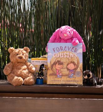 A children's book titled "Forever Cousins" is displayed outdoors on a wooden bench, surrounded by a plush teddy bear, a pink stuffed rabbit, a wooden toy, and a small raccoon plushie. Tall grasses serve as a backdrop.