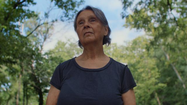 An elderly woman with short gray hair stands outside, looking thoughtfully into the distance. She's wearing a dark T-shirt and is surrounded by lush green trees under a partly cloudy sky.