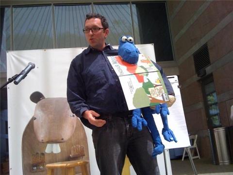 A man standing outside holds a blue puppet with an open book in its mouth. Beside him is a stage with a large animal backdrop. A microphone stand and a wooden stool are also visible. The setting appears to be a public event or performance.