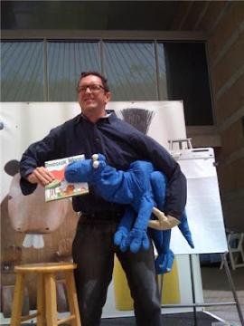 A person holding a blue dinosaur puppet in one arm and a children's book titled "Dinosaur Vs." in the other hand, standing on a stage with a stool and an easel in the background.