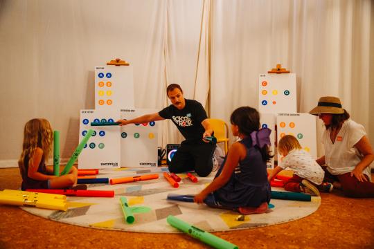 A man sits on the floor with a group of children, holding colorful foam tubes. Charts with symbols are in the background. The children, wearing casual clothes, listen attentively as they interact with the materials in a well-lit room.