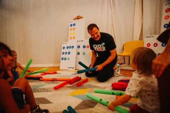 A man kneels on the floor, smiling, holding colorful tubes. He's surrounded by children, who also hold tubes. There's a chart with colored dots on a stand in the background. The atmosphere appears playful and educational.