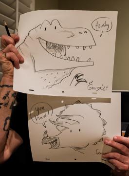 Two hand-drawn sketches are held up by a person. The top drawing shows a smiling dinosaur saying "Howdy." The bottom sketch depicts another dinosaur exclaiming "yee haw." The person has tattoos on their arms.