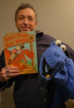 Man holding a book titled "Ridin' Dinos with Buck Bronco" and a blue dinosaur puppet. He is smiling at the camera.