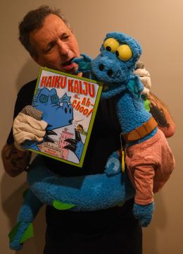 A person is holding a blue dragon puppet and a book titled "Haiku Kaiju & Ah-Choo!" The dragon is wearing a light brown shirt, and they are both holding microphones, appearing to perform together.