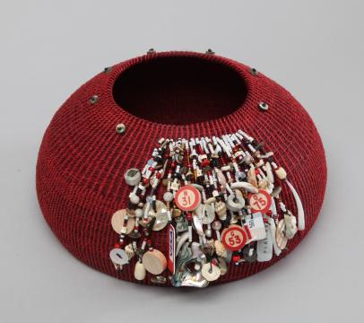 A round, red woven basket with a wide opening. It features an intricate decoration of colorful beads, shells, and small round tags with numbers on one side, creating a textured and vibrant design against the plain background.