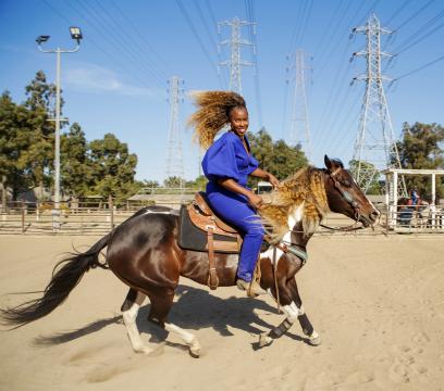 A person wearing a bright blue outfit rides a galloping horse in a fenced sandy arena. The rider's hair flows back dramatically. Power lines and towers are visible in the background under a clear blue sky.