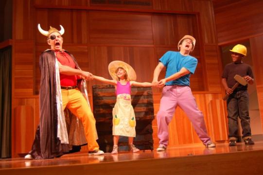 Four people on stage acting: One wears a Viking helmet and cape, another in a big hat and dress, a third in a casual outfit, and the last in a construction hat holding a tool, all appearing energetic and expressive.