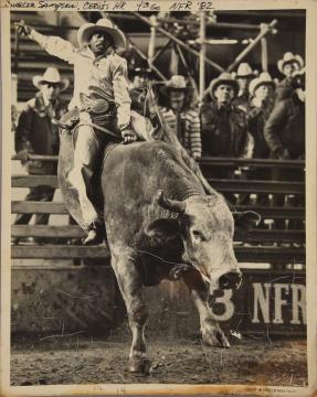 A rodeo rider in a cowboy hat and chaps rides a bucking bull in an arena, surrounded by spectators in cowboy hats. The number 3 and “NFR” are visible on the barrier. The photo is black and white and has handwritten notes at the top.