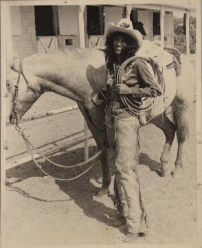 A smiling woman dressed in cowboy attire, including a hat and chaps, stands beside a saddled horse. She holds a coiled rope, with stables and open barn doors visible in the background.